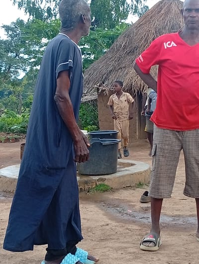 This image depicts an outdoor scene in Tse Igbudu, Nigeria, likely a rural or semi-rural setting. In the foreground, a man on the left wears a long, dark blue robe and light blue puffy sandals. To his right, a young boy in a light-colored shirt and pants stands near a large black bucket. Behind the boy, a man on the right is visible from the waist down, wearing a red t-shirt with white lettering and black and white checkered shorts. He is also wearing black and gray Adidas slides. Further in the background, a thatched-roof structure, possibly a hut, is present. The ground appears to be dirt and there are scattered green trees and foliage. The lighting suggests it is daytime, possibly overcast.