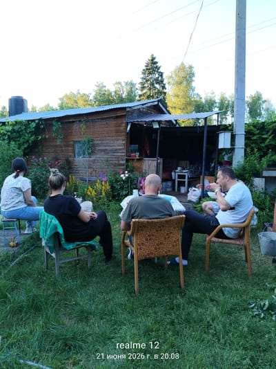 In the rural settlement of Космос-2 Снт, Russia, four individuals are gathered outdoors in a grassy yard, seemingly for a casual social event. The scene is dominated by a rustic wooden structure, likely a shed or outbuilding, with a corrugated metal roof. In front of this structure, a table is set up, around which three men and one woman are seated on chairs.

One woman, wearing a light-colored floral shirt and blue jeans, sits on a low stool on the left side of the frame. To her right, another woman, in a black t-shirt and dark pants, is seated on a wooden chair draped with a green cloth. In the center, a man in a green t-shirt is seated at the table, facing away from the camera. To his right, a man in a white t-shirt and patterned shorts is actively eating, holding a sandwich.

The environment suggests a warm day, with lush green grass covering the ground and trees forming a backdrop. There are various items scattered around, including buckets, bags of items, and gardening supplies, hinting at a lived-in, perhaps slightly cluttered, outdoor space. The overall impression is one of informal relaxation and social interaction in a rural setting.