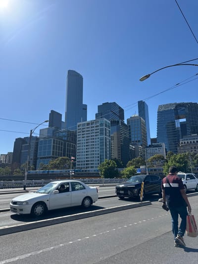 A cityscape is captured on a clear day, with various modern buildings reaching into the blue sky. A street runs alongside the buildings, with parked cars and a person walking with a KFC bag. Electrical lines and street lamps crisscross the sky. The buildings show diverse architectural styles, from tall, sleek towers to structures with intricate patterns.