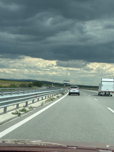 The image shows a view from inside a vehicle driving on a highway. The highway has multiple lanes and is bordered by a metal guardrail. Dark, heavy clouds are in the sky, with some lighter clouds visible. In the distance, there are rolling hills and some vegetation. Several vehicles are on the highway, including a silver SUV and a white truck. A billboard is visible in the distance on the left side of the highway. The scene appears to be in the rural area near Plodovitovo, Bulgaria, based on the provided context.