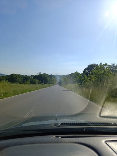 The image is a view from inside a car, looking out onto a straight asphalt road stretching towards the horizon under a clear, bright blue sky. The sun is shining brightly to the right, causing a lens flare. The road is flanked by green vegetation, including tall grass and trees. The location is near Svetlina, Bulgaria.