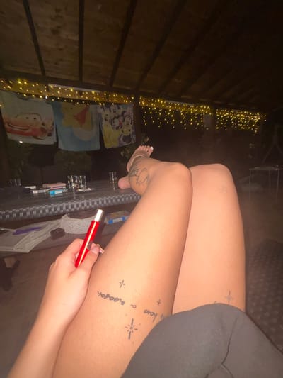A person is reclining in an outdoor, covered area at night. Their legs are crossed, and they are holding a red smoking device. Several tattoos are visible on their legs. A table in the background has drinks and what appear to be cigarette packs on it. String lights are draped across the ceiling of the covered area, and cartoon character-themed towels or blankets are hanging. The overall setting suggests a relaxed, social gathering in a backyard or patio space. There is no information within the image to suggest the location is in Viden, Bulgaria.