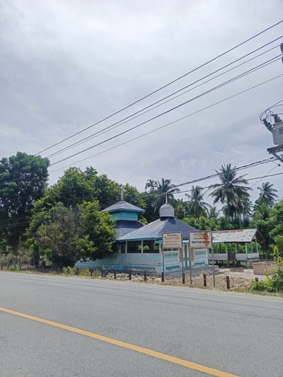 A street view shows the "Masjid Tuha Bugeng" (Bugeng Old Mosque) in Sawang, Indonesia, under a cloudy sky. The mosque features traditional blue and white architecture with a domed roof and an open veranda supported by posts. Two informational signs with Indonesian text are placed in front of the mosque, indicating "SITUS CAGAR BUDAYA" (Cultural Heritage Site). Adjacent to the mosque is an open-sided structure with a corrugated metal roof. Lush green trees, including palm trees, surround the buildings. Power lines stretch across the upper part of the frame, with an electrical transformer visible on a pole to the right. A paved road with a yellow center line runs in the foreground. The ground around the mosque is rocky and dry. There are no people visible in the image.