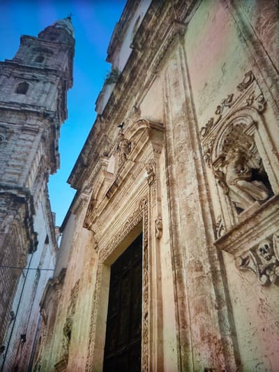 The image shows the exterior of a building, likely a church, with a tall, ornate tower visible on the left and a detailed facade on the right. The architecture appears to be historical, with stone construction and intricate carvings. The facade on the right features a prominent doorway and decorative elements, including a niche with a sculpture of a seated figure. The sky is a clear, bright blue. The perspective is from a low angle, looking upwards. The scene is devoid of any people or obvious activity. The stone exhibits weathering and discoloration.