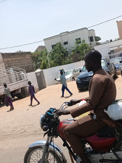 The scene depicts an unpaved street or open area in Hotoro, Nigeria, under a clear, bright sky. In the foreground, a dark-skinned male, dressed in a brown striped tunic and trousers, sits astride a red Bajaj Boxer 100 motorcycle. A white bag is secured to the rear of the motorcycle, and two plastic bottles, one white and one yellow, are visible near the fuel tank. His hands are on the handlebars.
In the mid-ground, from left to right, four other individuals are present. A light-colored pick-up truck with a white open-cage bed is parked on the far left. A young male in light clothing and purple trousers walks to the left, near the truck. Another young male, dressed in purple, walks towards the center-left. Further right, a male in a light blue tunic and dark blue trousers walks towards the left, balancing a yellow bowl on his head and holding a stick-like object in his right hand.
Behind these individuals, a concrete block wall and a white fence with a pillar marked '132' are visible. Several parked vehicles, including a dark sedan (possibly Mercedes-Benz) and lighter-colored sedans, are present on the right side of the frame. The background features a modern-style white building with dark windows and a flat roof, surrounded by green trees and vegetation.