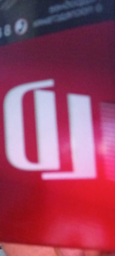 The image is a close-up, blurry, and poorly lit shot of what appears to be a red surface with large, white, stylized lettering. The lettering is abstract and appears to be two distinct shapes. One shape is a U-like curve on the left, and to its right is a vertical bar with a slight curve at the top. There is a pinkish-red glow emanating from the right side of the image. The background above the red surface is dark and blurry, with some faint circular and linear white shapes visible, possibly from a screen or other electronic device. The bottom of the image shows a blurry portion of what seems to be skin. The location of Vozdvijenka, Russia, is not discernible from the visual content of the image.