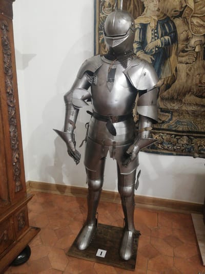 A full suit of polished metal plate armor stands upright on a dark wooden display base marked with a white "1" in an interior room of the Mir Castle Complex in Мірскі сельскі Савет, Belarus. The armor includes a closed helmet with a perforated visor, a breastplate, pauldrons, vambraces, gauntlets, faulds, tassets, cuisses, greaves, and sabatons, all secured with dark leather straps and buckles; chainmail is visible at the gorget. Directly behind the armor, a large, intricate tapestry depicting human figures in historical attire (one in blue and gold) and zoomorphic elements, such as lion heads, in muted blues, golds, browns, and creams, hangs on a light-colored wall. To the left, a tall, dark wooden furniture piece with detailed carved floral and vine motifs is partially visible, its base resting near the floor. The floor is covered with terracotta-colored hexagonal tiles, and a light-colored wooden baseboard lines the wall. The scene is illuminated by artificial light, casting subtle shadows on the wall.