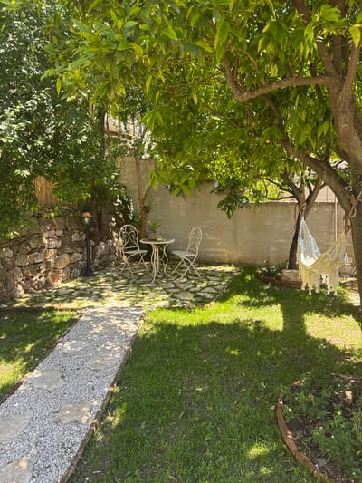 This outdoor scene, likely located in Baunei, Italy, features a garden setting on a sunny day. A tree occupies much of the foreground, with dense green foliage dominating the left side of the image. A white hammock is suspended between two tree branches in the background. A stone wall borders the garden on one side, while a gray concrete wall is visible further back. A white metal table and chairs sit near the wall, partially obscured by the tree. A gravel pathway with larger stones runs through the green grass lawn on the right side of the image. A circular planter sits along the edge of the path. The lighting suggests the scene is illuminated by natural sunlight.