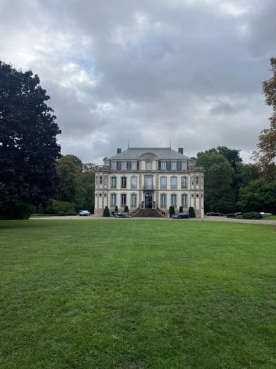 An external, eye-level, wide shot captures the Château Saint-Jean in Dorlisheim, France, under an overcast sky. The three-story, light-colored neoclassical building with a mansard roof stands centrally, flanked by dense green foliage. A wide, manicured green lawn occupies the foreground. A paved drive curves in front of the château, where multiple luxury automobiles are parked. On the left, a light-colored sports car is visible. Closer to the central entrance, a dark-colored high-performance vehicle is parked on each side of the main stone staircase. Further to the right along the drive, additional high-performance vehicles, including a red car and a white car, are parked. No individuals are visible. A large, dark-leaved tree frames the left side of the scene, while other deciduous trees with hints of autumn color line the right.