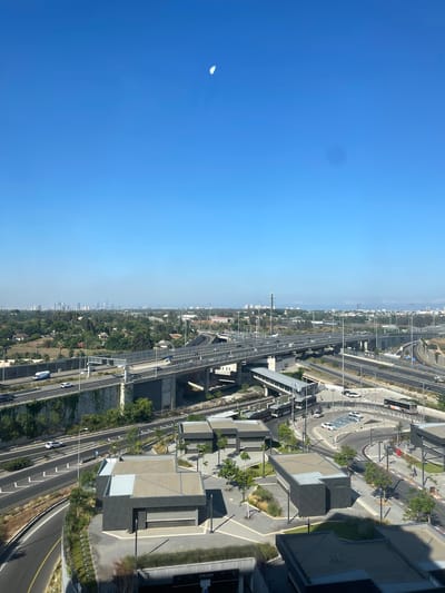 This is a high-angle, vertical shot of a modern city skyline in Ramot HaShavim, Israel, on a clear, sunny day. The left side of the image is dominated by a vast, bright blue sky, with a sliver of the crescent moon visible in the upper left quadrant. The right side of the frame showcases a bustling urban landscape, characterized by multi-lane highways, elevated train tracks, and contemporary architectural structures.

The scene depicts a dynamic intersection of transportation infrastructure and urban development. Cars and trucks are visible navigating the highways, indicating ongoing traffic flow. The train tracks appear to be part of a light rail system, with a station or platform visible amongst the buildings. The architecture consists of geometric, modern buildings with flat roofs and concrete facades, interspersed with greenery and landscaping. Some buildings have parking areas or plazas around them.

The time of day appears to be late morning or early afternoon, given the bright sunlight and the clear sky. The weather is fair and likely warm, typical of a sunny day in Israel. The distant cityscape suggests a sprawling urban area, with buildings stretching towards the horizon.

No people are clearly discernible in the image, but the presence of cars and infrastructure implies human activity and daily life. The overall impression is one of a well-developed, modern city with a strong emphasis on transportation and contemporary design. A sign with "PARK" is partially visible on a building on the far right.
