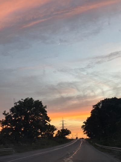 This is a photograph taken from a moving vehicle on a road in Tvardintsi, Bulgaria, during what appears to be sunset. The camera is oriented vertically, giving a unique perspective of the scene.
The sky dominates the left side of the image, transitioning from a pale blue and grey at the top to warm orange and yellow hues near the horizon, indicating the setting sun. Wispy clouds streak across the sky, with a sliver of the crescent moon visible in the upper left quadrant.
On the right side of the frame, a dark, curving asphalt road is visible, bordered by a white guardrail. Tall, dark green trees line the right side of the road, obscuring much of the background. In the distance, silhouetted against the bright sky, are power lines and what appear to be utility poles, suggesting a semi-rural or suburban setting.
The overall mood of the image is serene and contemplative, capturing the beauty of a natural sunset juxtaposed with man-made infrastructure. The lighting suggests it is late evening. There is no visible text in the image.