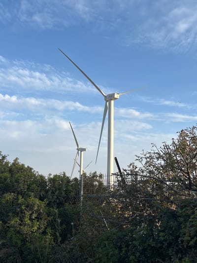 The image shows two wind turbines in Emek HaMaayanot Regional Council, Israel. The sky is blue with scattered white clouds. A tree and foliage partially obscures the view of the turbines and a metal fence is visible near the trees. The turbines are white, with three blades each. The logo "Aicon" is visible on one of the turbines.