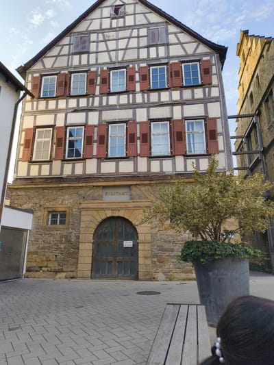 The image shows a half-timbered building in Neuenstadt am Kocher, Germany, likely the "Forstamt" based on the inscription above the arched doorway. The building has multiple windows with red shutters, some open and some closed. Below the half-timbered section is a stone facade with a large, arched wooden door. A tree in a large planter is situated in front of the building, and a wooden bench is in the foreground. The sky is partly cloudy. The scene appears to be a street or courtyard with cobblestone paving.