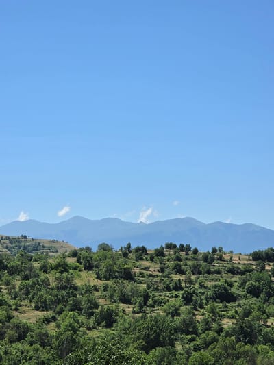This media file captures a serene landscape under a clear, bright sky. The scene is divided into two distinct parts: a vast expanse of blue sky on the left and a lush, green mountainside on the right. The mountainside is covered in dense vegetation, with trees and shrubs growing in abundance, suggesting a warm, sunny day. The terrain appears to be sloped, with evidence of terraces or natural contours formed by the vegetation.
In the distance, layers of mountains fade into a hazy blue, creating a sense of depth and scale. A few wispy clouds are visible against the sky, adding subtle texture to the otherwise uniform blue. The overall impression is one of natural beauty and tranquility. There are no people or man-made objects visible in the frame, emphasizing the untouched nature of the landscape. The sunlight suggests it is daytime, likely mid-morning to late afternoon, given the brightness of the sky and the clear visibility of the distant mountains. The location is identified as Slashten, Bulgaria, adding a geographical context to this picturesque view.