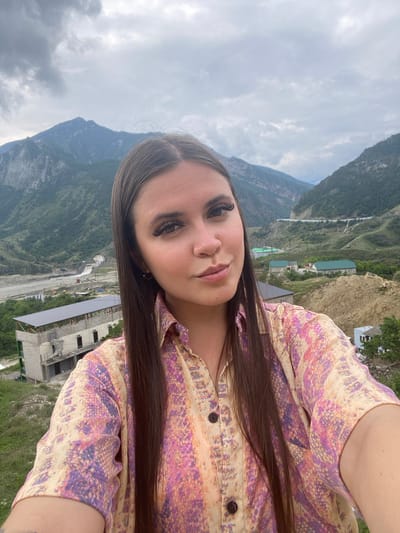A medium shot shows a young woman with long, dark hair and dramatic makeup taking a selfie in an outdoor environment. She is wearing a colorful, patterned shirt with shades of pink, purple, and yellow. Her expression is slightly pouty. In the background, a mountainous landscape is visible with some buildings and infrastructure, suggesting a semi-rural or developing area. The sky is overcast. The specific location of Chalda, Russia, is not identifiable from the visual elements of the image alone.