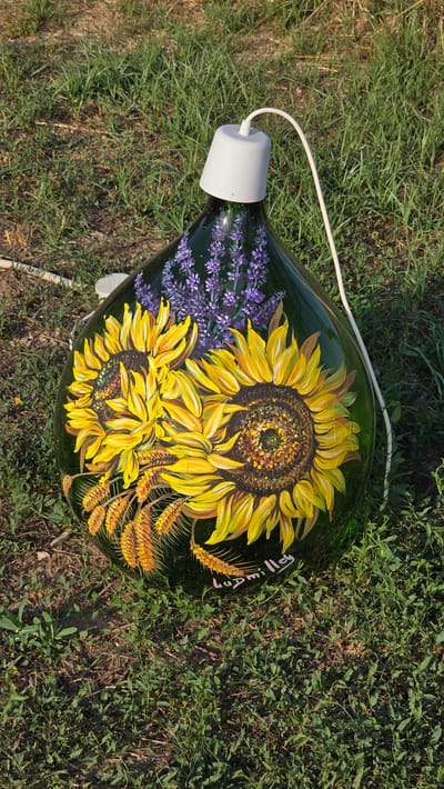 A green glass demijohn, painted with sunflowers, lavender, and wheat stalks, sits on grass. A white electrical cord emerges from a grey cap on the demijohn's opening. The demijohn is decorated with the name "Ludmilla" in pink cursive lettering. The scene is outdoors, with green grass covering the ground, suggesting a natural or garden setting. The image does not contain any specific visual cues to pinpoint a location within the city of Tuscania, Italy.