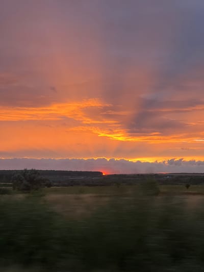 A landscape photograph captures a dramatic sunset over Kevdo-Mel'sitovo, Russia. The sky is ablaze with vibrant hues of orange, yellow, and pink, with rays of sunlight piercing through scattered clouds. A thin band of clouds borders the horizon, edged with a bright golden light. Below the sky, a silhouetted line of trees marks the transition to the middle ground, which appears as a blurred field. The foreground is also blurred, showing a dark green expanse, suggesting motion or a shallow depth of field. The sun itself is a small, red orb just above the horizon, partially obscured by the clouds. The overall impression is one of natural beauty and the ephemeral quality of a sunset.