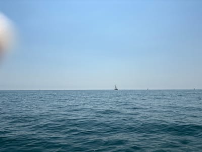 The image depicts a serene seascape with a vast expanse of calm, dark blue water stretching towards a clear, pale blue sky. The horizon line is sharp and distinct, dividing the water from the sky. Several sailboats are visible in the distance, scattered across the water, with their sails catching the light. One sailboat, positioned slightly off-center, is more prominent than the others. The water exhibits gentle ripples, indicating a light breeze. The overall atmosphere is peaceful and tranquil, characteristic of a pleasant day at sea. While the image is identified as being in Cesenatico, Italy, this specific viewpoint does not offer any recognizable landmarks to confirm the location within the city.
