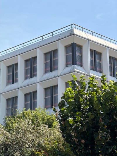 The image shows a close-up, low-angle shot of a modern building with a white facade and a series of dark horizontal beams visible through its windows. The windows appear to be covered with a plastic sheeting. The building's architecture is characterized by its repetitive geometric design, with angled frames around each window. A metal railing runs along the edge of the building's exterior. To the right of the building, lush green foliage from trees and bushes is visible, partially obscuring some of the building's features. The sky is a clear, pale blue, suggesting it is daytime with good weather. The overall scene is one of urban architecture juxtaposed with natural elements. There is no visible text in the image. The location context provided is Rungis, France.