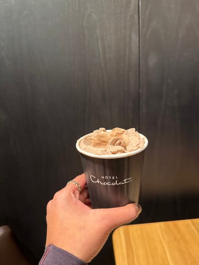 A close-up image depicts a human left hand holding a dark-colored paper cup, emblazoned with the white text "HOTEL Chocolat." The cup contains a light brown, semi-frozen substance, likely a chocolate dessert, featuring textured peaks and swirls on its surface. The hand displays a silver-toned ring with an interlocking circular or oval pattern on the ring finger, and dark-painted fingernails. A dark fabric sleeve is visible around the wrist. The background features a dark, vertically paneled wooden wall occupying the majority of the frame, with a light-colored wooden table surface visible in the lower right corner. The scene is situated within a Hotel Chocolat establishment, specifically at the Merry Hill Shopping Centre in Brierley Hill, Dudley, United Kingdom.