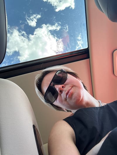 The image is a selfie taken from a low angle, with the subject's head tilted downwards. The subject is a person wearing sunglasses and a black top, with blonde hair. They are wearing jewelry on their wrist and earrings. The background shows a bright blue sky with fluffy white clouds, suggesting it is daytime and the weather is fair. The setting appears to be inside a car, with parts of the car's interior visible, including the ceiling and upholstery. The orientation of the image is upside down, as indicated by the way the sky and the car's interior are positioned. There is no visible text in the image.