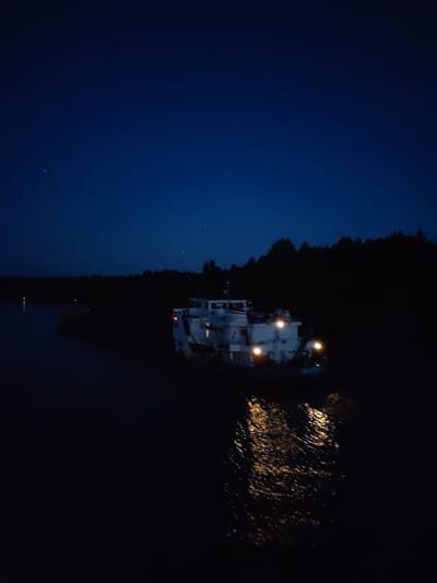 A low-angle nighttime shot of a white passenger ship on a body of water, likely a river or a bay. The ship is illuminated with several lights, which reflect in the water's surface below. The surrounding area consists of a dark treeline. The night sky above is dark blue with a few visible stars. There is no evidence to link this picture to Uzhla, Russia.