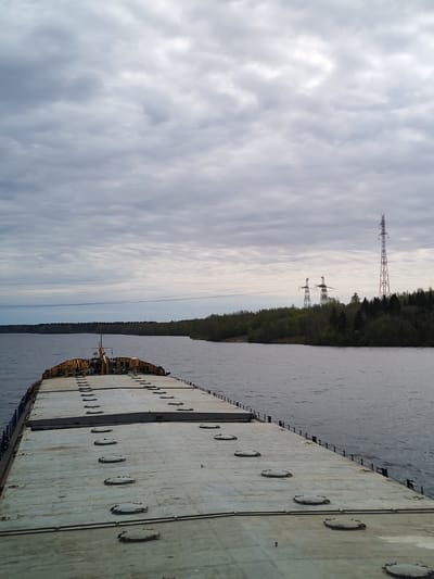 The image presents a view from the deck of a barge on the water, possibly a river or large lake. The deck of the barge is gray and featureless except for multiple circular hatches. The water is gray and slightly choppy. In the distance, a forested shoreline is visible. Three power transmission towers are visible along the shoreline. The sky is overcast with gray and white clouds.

While it is not possible to pinpoint the exact location as Борисово, Russia using just this image, the presence of a wide body of water with forested shores and power transmission towers aligns with the characteristics of rivers and lakes in Russia.