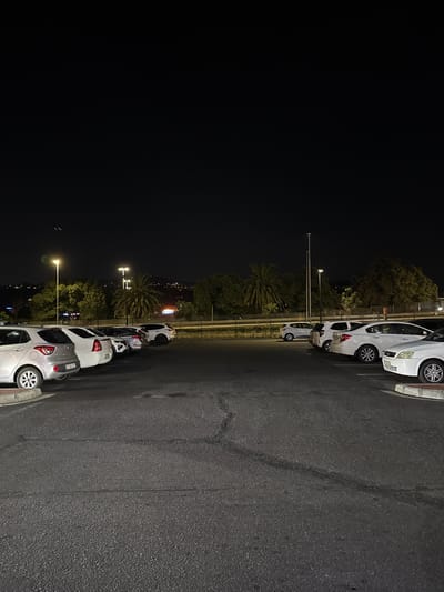 The image depicts a nighttime scene in Bellville, South Africa. Several cars are parked along a street illuminated by streetlights. Palm trees and other vegetation are visible between the cars and the darker background, where city lights are also present. The road surface appears to be asphalt. There are no people visible in the image.