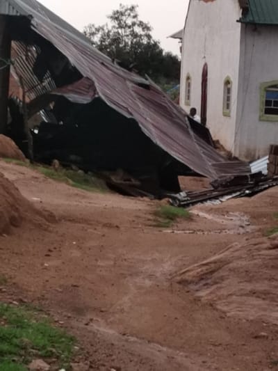 The scene depicts a collapsed corrugated metal roof of a building, lying in a pile on the ground in front of a white building with a green roof, likely a church or community center. The roof is heavily damaged, with multiple sections broken and displaced. The building is located at coordinates latitude 9.93, longitude 8.86 in the city of Gerin Gada, Nigeria. The ground is dirt and appears damp, possibly from recent rain. There are no people visible in the image. The scene suggests a recent natural disaster like a storm or wind event.