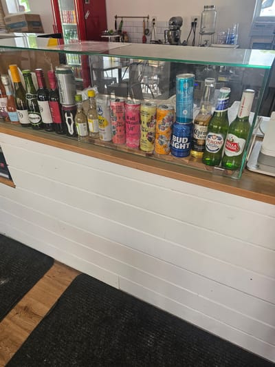 The image shows a refrigerated display case filled with alcoholic beverages, including beer, wine, and canned cocktails. Brands visible include Stella Artois, Peroni, Corona Extra, Bud Light, Two Chicks, Cali Cocktails, Dark Horse, and several wine varietals such as Pinot Grigio, Rosato, and Syrah. The display case appears to be located in a store, potentially a liquor store or market.  The flooring is dark, possibly carpet, adjacent to white paneled walls. There is no visual evidence to confirm the location is Key Largo, United States of America.