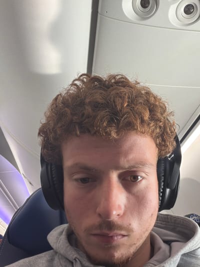 A close-up, candid portrait of a young man is captured within an airplane cabin. The man, positioned in the lower right of the frame, has light brown, curly hair and light stubble on his chin and upper lip. He wears black over-ear headphones and a grey hooded sweatshirt. His eyes are open, and his gaze is directed towards the viewer, showing a neutral or slightly thoughtful expression. His visible left eye is light brown.

The setting is distinctly an airplane interior, evident from the white overhead panel in the upper left, featuring two circular reading lights and what appear to be air vents. A dark blue airplane seat is visible in the lower right corner, and below it, part of the cabin interior shows a purple-hued ambient light, possibly from mood lighting or a window reflection. The lighting in the cabin is bright and even. On the grey hoodie, a faint and indistinct logo or text is partially visible on the upper right shoulder area, but its specific content cannot be clearly discerned. The overall impression is a personal moment during air travel.