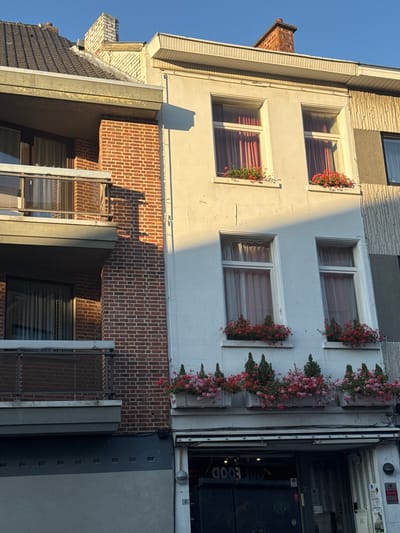 The image shows the facade of a building in Strombeek-Bever, Belgium. The building has a brick exterior on the left side, with a balcony. To the right of the brick section is a white-painted section of the building with several windows adorned with flower boxes. The windows have red curtains. Below the white section is a storefront with a sign that appears to read "HOT FOOD". The sky is clear and blue, and sunlight casts shadows on the building.
