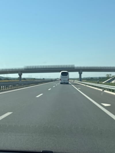 The image shows a highway with a bus driving away from the viewer. A bridge is visible in the background, spanning across the highway. The sky is clear and blue. The highway appears to be in a rural area, with some vegetation visible on the sides. The location is the E75 highway near Jarak, Serbia, on a sunny day.