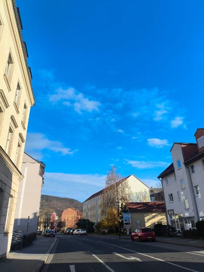 The image depicts a street scene in Freital, Germany, under a clear, bright blue sky with sparse white clouds. The perspective is from the center of the asphalt road, looking forward and slightly uphill.

On the left, a multi-story building with a light beige facade and numerous windows occupies the foreground, extending upwards and out of frame. Further down the street on the left, a pale grey building is partially visible, followed by parked vehicles lining the curb. In the mid-ground, a reddish-brown building with a prominent cross on its facade, likely a church or similar institution, stands at a crossroad. Hills covered with bare trees are visible in the background.

On the right side of the road, a light grey, multi-story building with a red-tiled gable roof and multiple windows is present in the foreground. A white business sign is affixed to the right side of the building. Further down the street on the right, a long, light yellow multi-story building with a red-tiled roof is visible. A smaller, single-story building with a red roof is situated between these two structures. Numerous vehicles are parked along both sides of the street. A red hatchback is prominently parked on the right side of the road, facing away from the viewer.

Bare deciduous trees, indicative of late autumn or winter, are scattered throughout the scene. Streetlights are visible. No individuals are discernible within the frame. The overall impression is a quiet urban street on a sunny day.