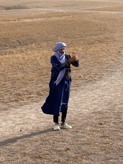 A person in traditional Moroccan attire stands in a field, holding a mobile phone and taking a picture. The person is wearing a blue robe with gold embellishments, black pants, white shoes, and a light blue head covering. The field is dry and grassy, with a dirt path visible. The location is in Sidi Badhaj, Morocco.