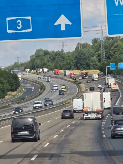 The image depicts a multi-lane highway in Montabaur, Germany, on a sunny day with scattered clouds. The foreground features a black Mercedes-Benz V-Class van with license plate "DAU US 666" driving in the right lane. Behind it, in the left lane, is a dark-colored convertible. Further ahead, a white Mercedes-Benz Sprinter van is visible, followed by several trucks and other vehicles.
Overhead, a large blue highway sign indicates "3" on the left, and an upward-pointing arrow. To the right of this sign, another blue sign displays destinations including "Frankfurt/Oder" and "Wiesbaden." In the background, a dense line of green trees borders the highway, and several tall electricity pylons are visible against the sky. The road surface appears to be asphalt, with clear lane markings. The overall impression is one of active traffic flow on a major German highway.