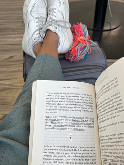 The image depicts a person sitting on a chair with their legs resting on a piece of luggage. The person is wearing white sneakers and teal blue pants. A bright pink, blue, and orange tassel hangs off the side of the luggage. The person is reading a book titled "The Power of Now," the open pages of which are visible in the image. The book is lying on their lap. The image is taken indoors, likely at an airport or terminal, as a wooden floor can be seen in the background. The location of the image, based on the provided coordinates, is within the city of Tlajomulco de Zúñiga, Mexico.