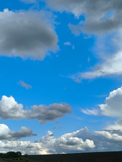 A panoramic view of a bright blue sky filled with a mix of fluffy white and darker gray clouds, indicative of fair weather with potential for passing showers. The horizon is low in the frame, showing a dark, flat expanse that could be a field or undeveloped land, with a line of dark green trees visible on the far left. The image appears to be taken outdoors, possibly during the daytime, in an open rural or semi-rural setting, consistent with the outskirts of a location such as Bol'shie Ploty, Russia. There are no people or animals visible in the frame.