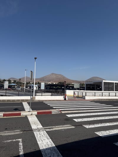 The image is taken in San Bartolomé, Spain, and shows a street scene with a clear blue sky overhead. A mountain range can be seen in the distance, partially obscured by haze. A modern, white building with dark horizontal slats is located near a pedestrian crosswalk, which is marked with thick, white stripes on the asphalt road. A red and white striped pole stands at the edge of the crosswalk. Street lights are visible along the road, and palm trees can be seen in the background, lining the street. Several parked cars are visible.