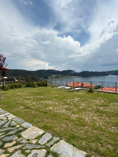 The image shows a view of Dospat Dam in Dospat, Bulgaria. In the foreground, there is a stone pathway bordering a grassy lawn. Beyond the lawn, a black metal fence separates it from a tiled roof structure and several lounge chairs. The dam itself is visible in the middle ground, surrounded by a dense forest and rolling hills. The sky is filled with dramatic, cloudy formations.