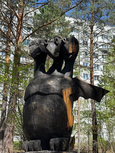 The image depicts a large, dark, wooden sculpture in an outdoor setting. The sculpture appears to be of a dragon or mythical creature, with visible wings, a stout body, and multiple faces with prominent eye holes. The wood has a charred or burned appearance, adding to its unique texture and aesthetic.
The sculpture is positioned in front of a backdrop of trees and a building with multiple windows, suggesting an urban park or landscaped area. Based on the location in Sayansk, Russia, it's likely situated in a public space like a park or recreational area. The environment includes both deciduous and coniferous trees, indicating a temperate climate. The branches and leaves suggest it is spring or summer. The presence of a building with modern architecture suggests the sculpture is located within or near a residential or commercial area of the city.