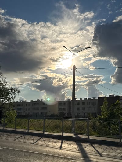 A street lamp with two lights stands in the foreground, silhouetted against a dramatic sky filled with dark, heavy clouds and bright sunbeams. The sun is partially obscured by clouds, creating a bright halo effect. Below the lamp, a multi-story building with rows of windows stretches across the mid-ground. A metal fence with a diamond pattern runs in front of the building. The ground in the foreground is a paved road with a crosswalk, casting long, geometric shadows from the fence onto its surface. The scene appears to be a street in the city of Rastunovo, Russia, during daytime.