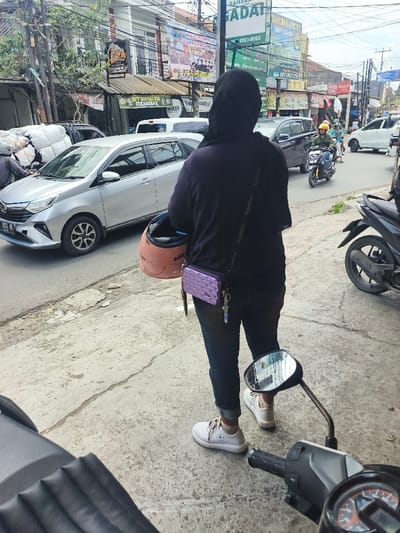 A person wearing a black hijab, dark shirt, blue jeans, and white sneakers stands on a sidewalk. They hold a pink helmet and have a small purple purse on their shoulder. A silver car and several motorbikes are on the road behind them. A black motorbike is parked in front of the person, partially blocking the view. Buildings and billboards line the street.