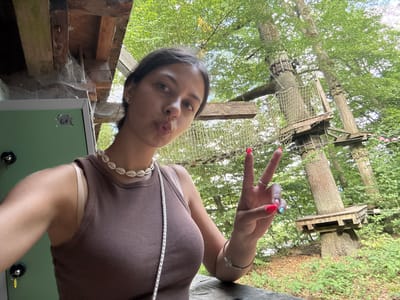 A young woman with dark hair is taking a selfie in an outdoor adventure park. She is wearing a brown tank top and a shell necklace. She is making a peace sign with her right hand, which has bright pink nail polish. In the background, there are trees with climbing structures and nets, typical of a high ropes course. To the left, a green locker is visible with the number 61 on it. The scene appears to be set in a natural environment with trees and foliage. This location is identified as Seulberg, Germany.