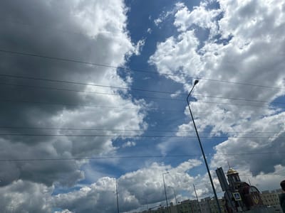 This image, taken in Stroykeramika, Russia, captures a cloudy sky with a mix of dark and light clouds. Several power lines are visible in the foreground, stretching horizontally across the frame. A tall street lamp stands prominently on the right side of the image. In the background, a building with a golden dome, resembling a church, is visible. Several other buildings and street lamps are also present. The overall scene appears to be an outdoor urban environment under a dynamic sky.