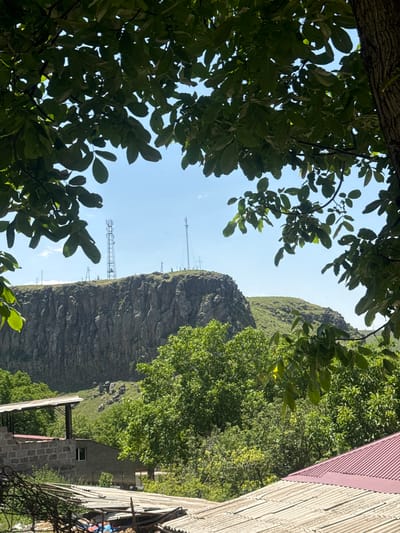 This is a photograph taken in the city of Bjni, Armenia. The scene is framed by the foliage of a large tree in the foreground, with its branches and leaves obscuring parts of the view. Beyond the trees, a rugged, rocky cliff face dominates the middle ground. Two tall radio towers stand atop this cliff. The sky above is clear and blue, with a few wispy clouds. In the lower portion of the image, there are several residential structures, including buildings with brick and concrete walls, covered by corrugated metal roofs. The environment is predominantly natural, with lush green vegetation and a prominent geological formation.