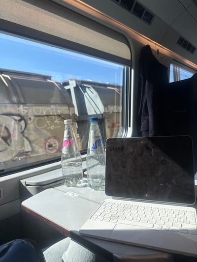 The image shows an interior view, likely inside a train car, in Sion, Switzerland. A window reveals a view of the outside, including graffiti on a building. On a table in front of the window are two water bottles, an iPad, and a white keyboard. A dark-colored garment is placed on the upper-left area of the frame, suggesting an article of clothing. The image is oriented in a non-standard way, with the top of the train car on the left.