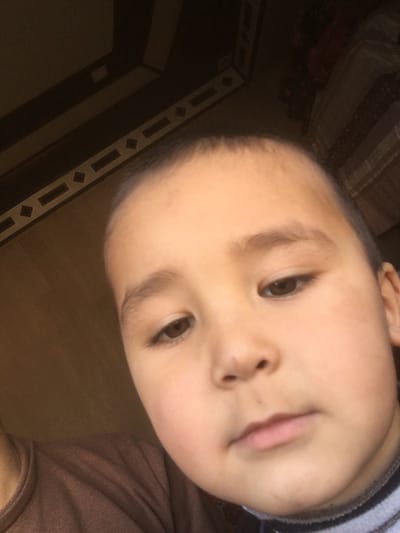 A close-up, indoor image depicts a young child, likely male, with fair skin, dark brown eyes, and short dark hair, facing slightly downwards and to the viewer's right. The child's face occupies the majority of the frame, capturing the forehead, eyes, nose, and mouth, with a neutral expression. The child wears a brown or dark beige long-sleeved garment, with a striped collar (white and dark horizontal stripes) visible underneath. To the upper left of the child, a dark floor or wall surface features a decorative border composed of repeating light-colored rectangular and diamond shapes. To the upper right, a dark, patterned fabric, possibly part of a bed or piece of furniture, is visible and out of focus. The lighting suggests an indoor setting. The image depicts an indoor environment. The specific location within the city of Elatan, Uzbekistan, beyond a residential interior, is not discernable from the visual evidence.