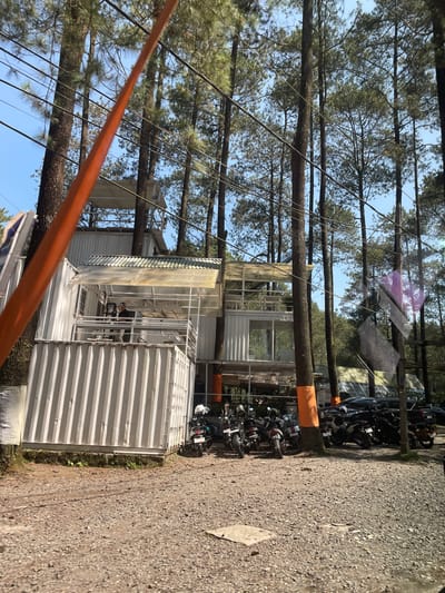 The image depicts a multi-level structure constructed primarily from white corrugated shipping containers, set amidst a dense pine forest. This location is identified as Cikole, Indonesia.

The building features multiple stories with balconies and large windows. The top levels have transparent or translucent roofing elements, possibly awnings or skylights. On a mid-level balcony on the left side of the structure, at least two individuals are visible. One person, wearing a dark cap and shirt, stands near a railing.

Numerous tall pine trees dominate the background and surround the building, with some tree trunks integrated into the structure's design or piercing through its levels. Several tree trunks in the foreground and near the building are wrapped with orange fabric or tape at their bases. Overhead, numerous black power lines crisscross the blue sky.

The ground is covered with light brown gravel. To the right of the building, a collection of motorcycles of various makes and models are parked. Further right and in the background, several dark-colored cars are also parked. An prominent orange, fuzzy strap or cable runs diagonally from the upper left to the mid-right in the immediate foreground, partially obscuring the view. The scene is illuminated by bright daylight, indicating clear weather conditions. Reflections are visible on the right side of the image, suggesting the photograph was taken through a window.