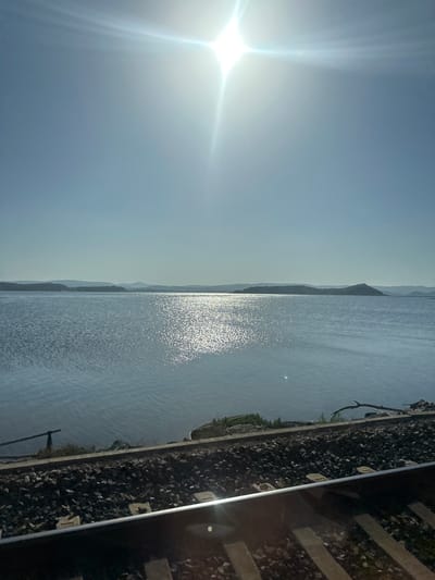 The image depicts a scenic view of a body of water, likely a lake or a wide river, bordered by a railway track on one side and distant hills or mountains on the other. The sun is prominent in the sky, casting a bright glare and creating shimmering reflections on the water's surface. The sky is a clear blue, suggesting fair weather. The railway track, made of concrete sleepers and ballast, runs parallel to the water. A few sparse trees and vegetation are visible along the edge of the water near the tracks. The overall impression is one of tranquility and natural beauty, possibly captured during a bright, sunny day. The time of day appears to be late morning or early afternoon, given the sun's position and brightness. There is no visible text in the image.